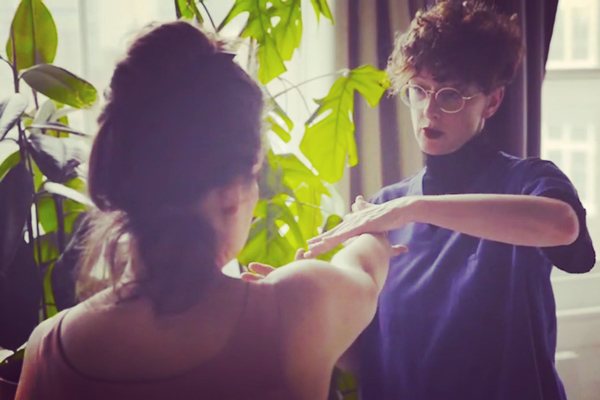 Two people interacting indoors, one person with short curly hair wearing glasses is demonstrating something with their hands to another person with long hair, in a setting filled with indoor plants. The image conveys a sense of connection and learning in a nurturing environment.