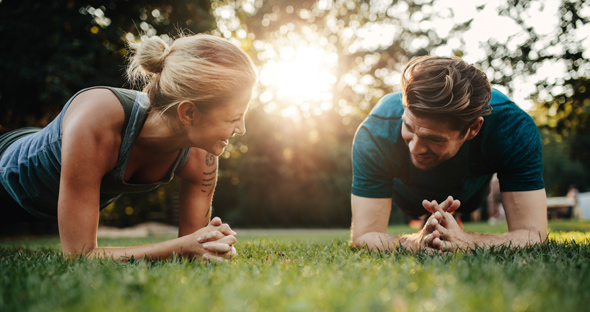 A man and woman engaging in a plank exercise outdoors during sunset, smiling and supporting each other on a grassy field. Fitness motivation and teamwork in a natural setting.