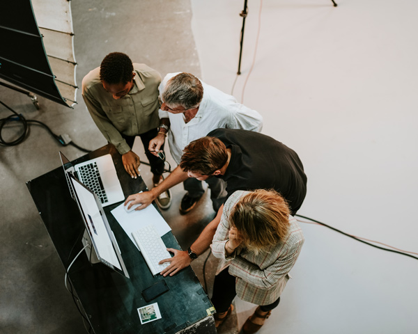 A diverse group of four individuals collaborating and discussing work over a laptop on a table in a creative workspace.