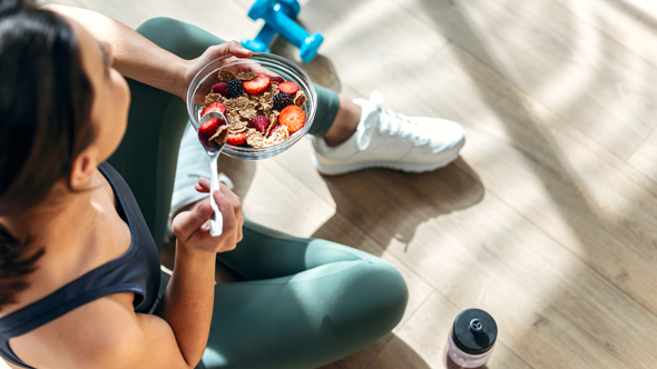 A person sitting on the floor enjoying a bowl of healthy granola topped with strawberries and raspberries, with a dumbbell and water bottle nearby.