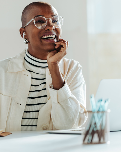 Smiling woman with short hair wearing glasses and earbuds, dressed in a striped sweater and light jacket, seated at a desk with a laptop and stationery.