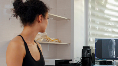 A woman with curly hair sits in a studio, looking at a computer screen displaying an X-ray image, with a spine model visible on a shelf nearby.