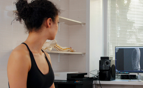 A woman in athletic wear sits in a medical office, facing away from the camera, while a large monitor displayed nearby shows an X-ray image of a spine, emphasizing health and wellness in the treatment environment.