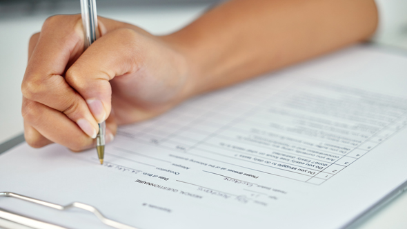 A hand writing on a document with a pen, focusing on filling out a form.