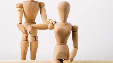 Two wooden mannequin figures, one standing and assisting the other that is seated, in a neutral backdrop.