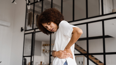 A woman with curly hair stands in a modern home, experiencing back pain while holding her lower back.