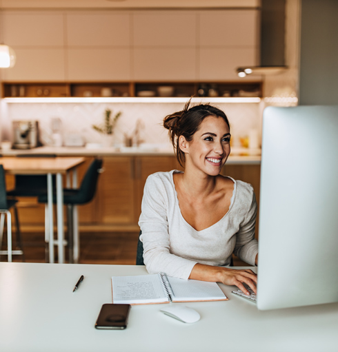 Smiling woman working on a computer at a modern kitchen table, with a notebook and smartphone nearby.