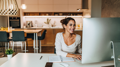 Woman working on a computer in a modern kitchen, smiling while typing. The scene conveys a productive home office environment.