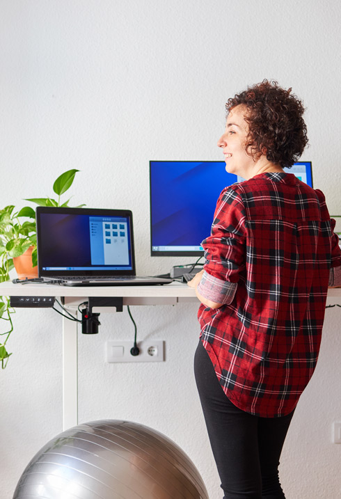 A person with curly hair stands at a standing desk, smiling while looking at two computer monitors. A green plant is on the desk, and a fitness ball is on the floor.