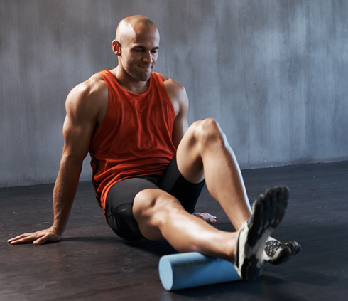 A man in a red tank top using a foam roller on his leg while seated on a gym floor, focusing on muscle recovery and flexibility.