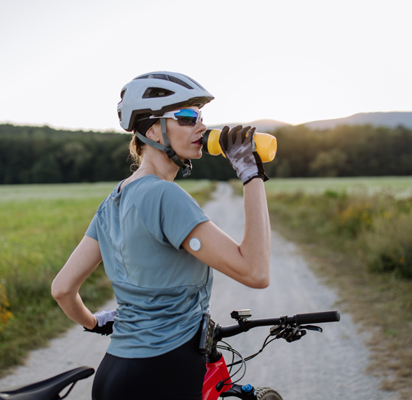 A woman in a cycling outfit takes a drink from a yellow water bottle while standing next to her mountain bike on a dirt path, surrounded by a green landscape at sunset.