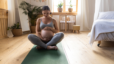 pregnant woman practicing yoga in a peaceful indoor setting, sitting on a green yoga mat with hands on her belly, surrounded by plants and natural light