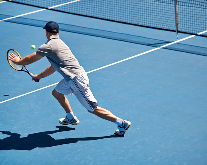 A man wearing a gray shirt and shorts is lunging to hit a tennis ball on a blue court, showcasing his athleticism and focus during a tennis match.