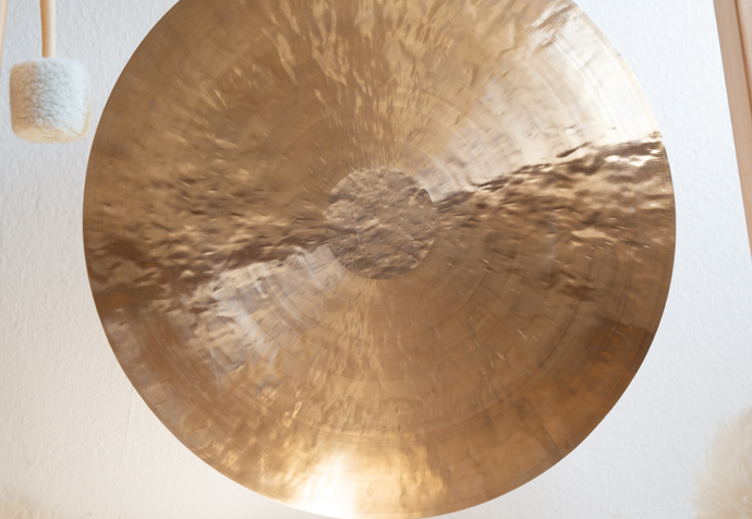 Large, shiny brass cymbal hanging on a wall with a mallet nearby, reflecting light and showcasing its textured surface. Ideal for music enthusiasts.