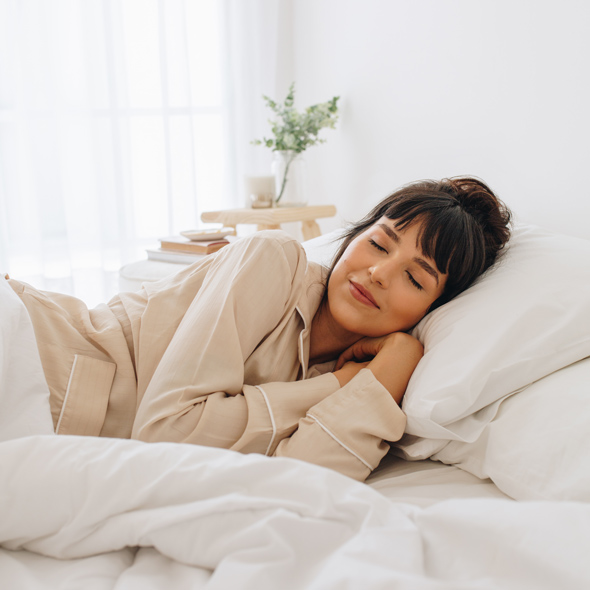 Young woman peacefully sleeping in a cozy bed, dressed in light pajamas, with a serene expression, surrounded by soft pillows and blankets. The atmosphere suggests relaxation and comfort.