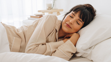 A woman in a cozy bed resting peacefully with a serene expression, wearing light-colored pajamas, surrounded by soft white pillows.