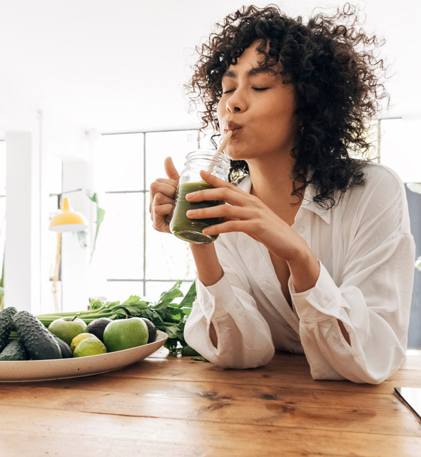 Woman enjoying a green smoothie in a bright kitchen, surrounded by fresh fruits and vegetables on a wooden table. Healthy lifestyle and green diet concept.