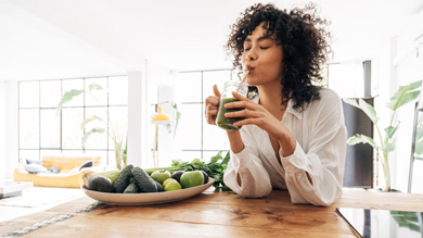 Woman enjoying a green smoothie while sitting at a wooden table, surrounded by fresh fruits and vegetables in a bright, airy kitchen. Healthy lifestyle.