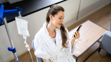 A woman in a white bathrobe sitting on a chair, smiling while looking at her smartphone, with an IV stand in the background, suggesting a comfortable recovery environment.