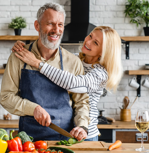 A smiling couple in a modern kitchen, with the man chopping vegetables and the woman playfully embracing him, surrounded by fresh produce including peppers and cucumbers. The scene reflects a joyful cooking experience and healthy lifestyle.