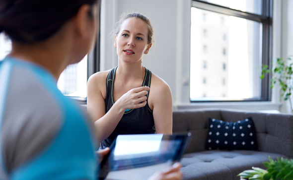 Two women engaging in a conversation in a modern indoor setting, one woman is listening intently while the other holds a tablet. The first woman appears to be in activewear, suggesting a focus on fitness or health coaching.