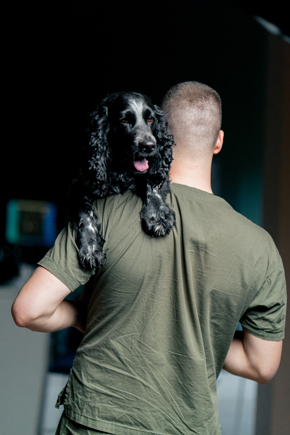 man in a green shirt holding a black and white dog on his shoulder, indoor setting, pet care