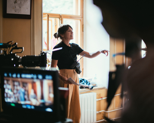 A woman in a black shirt and beige pants is gesturing while standing in a well-lit room, surrounded by film equipment. The focus is on her as she communicates, illustrating the dynamics of film production.