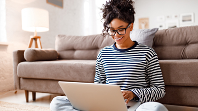 Young woman sitting on the floor using a laptop in a cozy living room, wearing glasses and a striped sweater, with a sofa and lamp in the background. Ideal for home office inspiration.