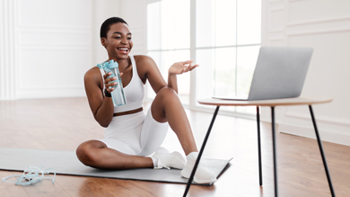 A woman in a white workout outfit smiling and sitting on a yoga mat, holding a water bottle while engaging in a virtual fitness session on her laptop.