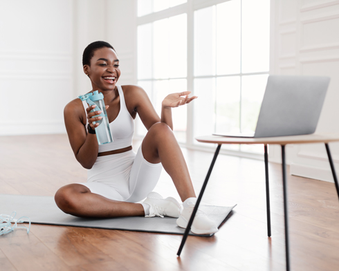 smiling woman in workout attire sitting on yoga mat, holding a water bottle and engaging with a laptop during a virtual fitness class