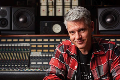 Man in a red plaid shirt smiling in a recording studio with audio equipment and speakers in the background.