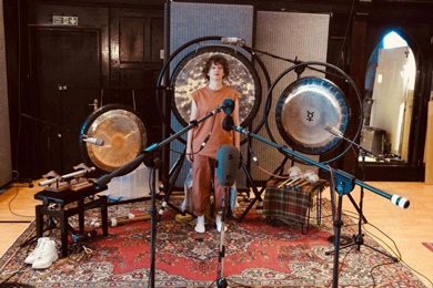 A person standing in a music studio surrounded by gongs, with microphones set up for recording, creating an atmosphere of sound healing and musical experimentation.