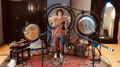 A person stands in a music studio surrounded by large gongs and other percussion instruments, with microphones set up for recording.