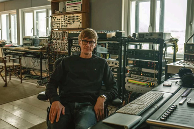 A person sitting in a music studio filled with electronic equipment and synthesizers, showcasing a creative workspace.